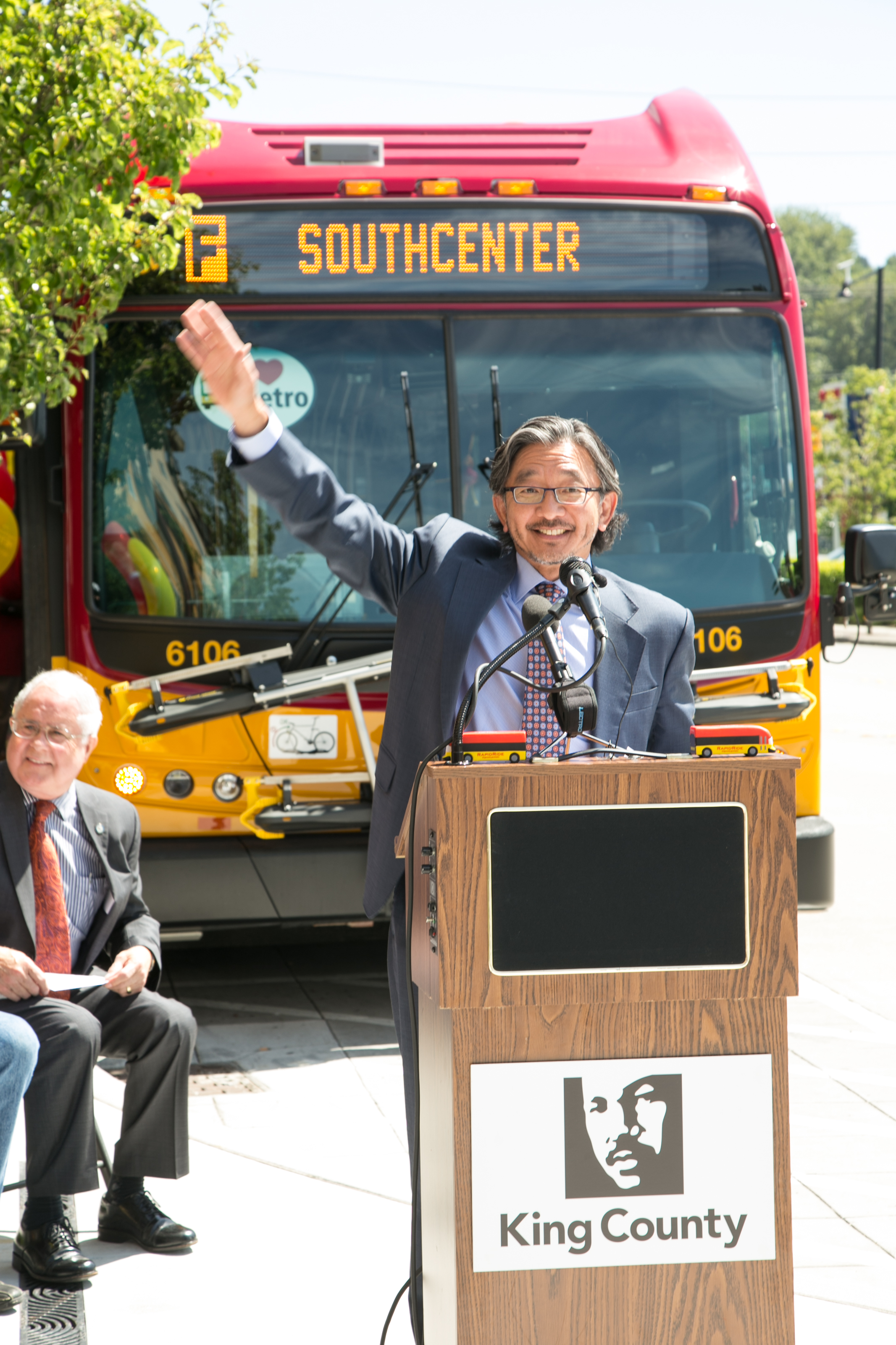 With a RapidRide F line bus in the background, Department of Transportation Director Harold Taniguchi waves.