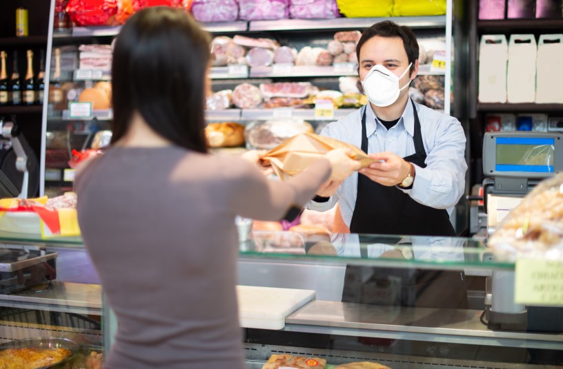 A clerk wearing a black apron and a face mask on handing an item to a woman in a shop.