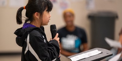 Girl speaks into microphone in front of crowd.