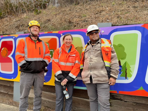 Three people stand in front of the "Welcome to Skyway" mural in Skyway, Washington