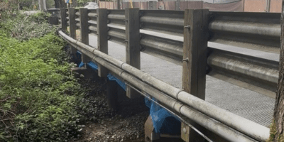 A bridge with guardrails over a creek. In the background is greenery and a building.