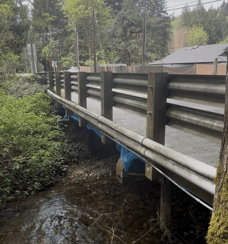 A bridge with guardrails over a creek. In the background is greenery and a building.