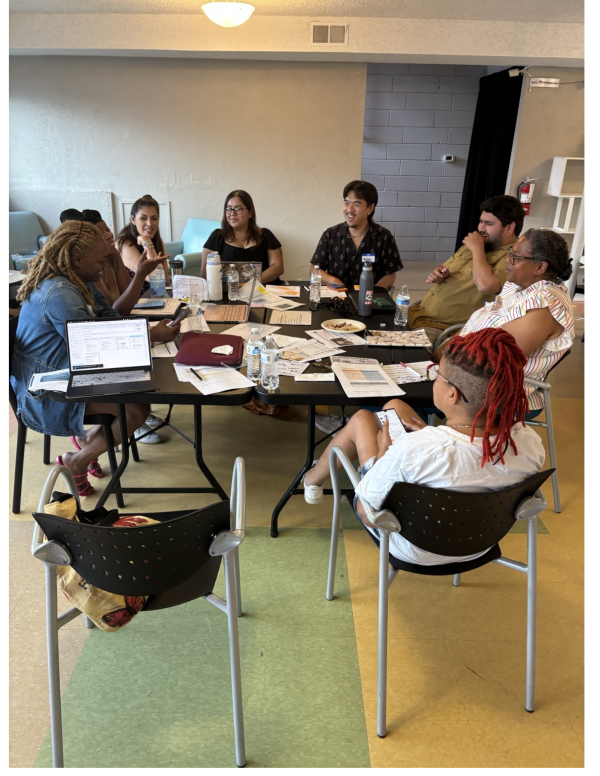 A group of people known as the Community Advisory Board discuss and laugh while sat at a table. On the table are flyers, nametags, and paper plates that had food on them.