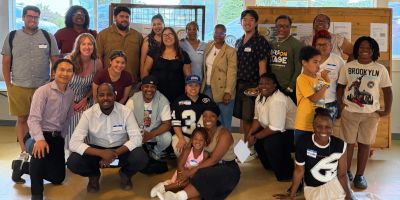 A group of people known as the Community Advisory Board for King County's Participatory Budgeting program pose for a picture in a room. In the background is a window and wooden bulletin board.
