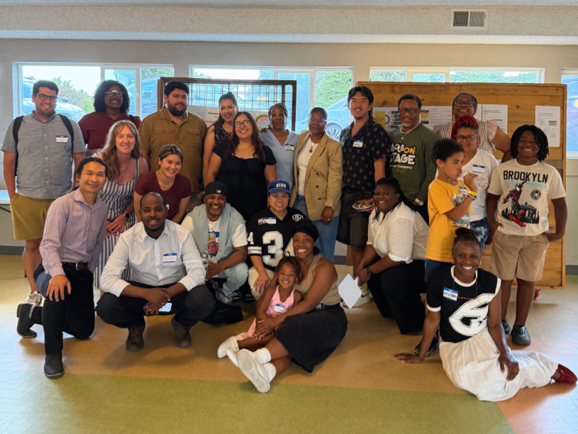 A group of people known as the Community Advisory Board for King County's Participatory Budgeting program pose for a picture in a room. In the background is a window and wooden bulletin board.
