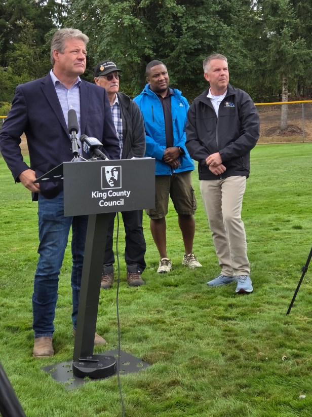 A man looks out to the crowd while standing behind a microphone on a podium featuring the King County logo with Martin Luther King's image. Behind him are three adults who are also looking to the crowd. They are standing on grass, with trees and a fence behind them.