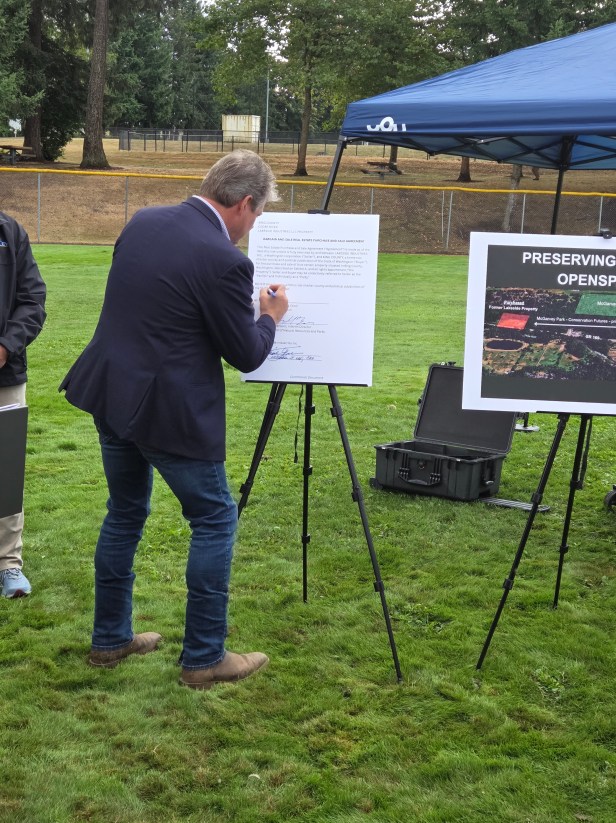 A man signs a posterboard-sized agreement positioned on an easel at Fairwood's 60th Anniversary Celebration. Another posterboard is on an easel nearby. The easels are positioned on grass.