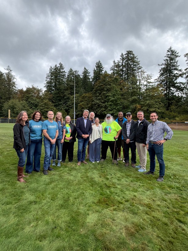 A group of adults pose for a picture while standing on open grass and trees behind them.