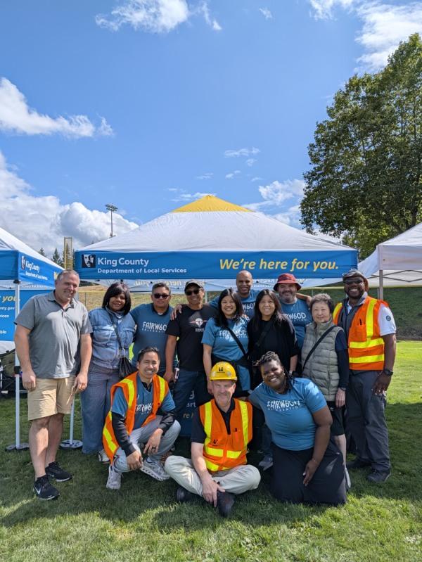 An adult group of King County employees pose for a group picture at Fairwood's 60th Anniversary Celebration on Saturday, Aug. 29, 2026. They are located in a grassy area, and behind them is a department-branded tent and trees.