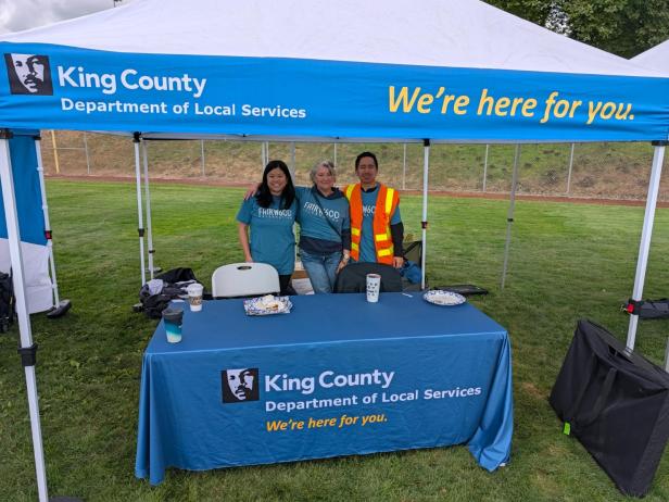 A trio of county employees pose for a picture while standing under a department-branded tent at Fairwood's 60th Anniversary Celebration on Saturday, Aug. 29, 2026. In front of them is a table with a department-branded tablecloth, and they are in a grassy area.