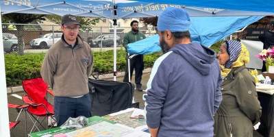 A King County staff member speaks to residents while standing under a branded canopy tent and behind a table with maps, fliers and other information on it.
