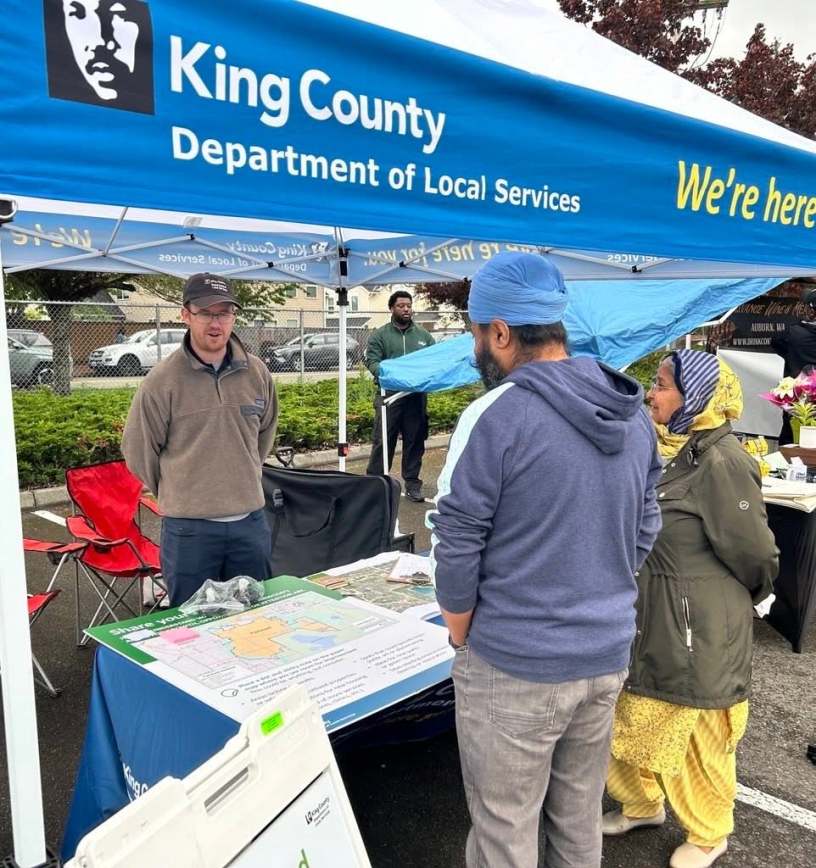 A King County staff member speaks to residents while standing under a branded canopy tent and behind a table with maps, fliers and other information on it.