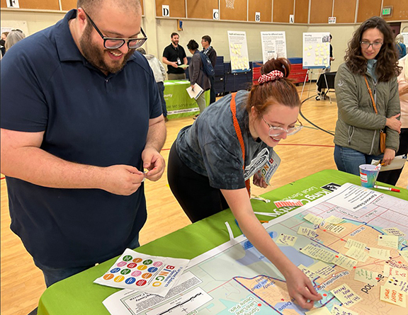 Two people place sticky notes on a map of the community of Fairwood in King County, Washington. The map is set atop a green table. Next to them is a person who is observing. In the background are people at other stations.