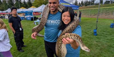 Two adults pose wearing t-shirts celebrating Fairwood's 60th anniversary pose for a picture with a large snake around their shoulders. They are standing in an open fields, and in the background are other people, tents, trees, and gloomy sky with gray clouds.