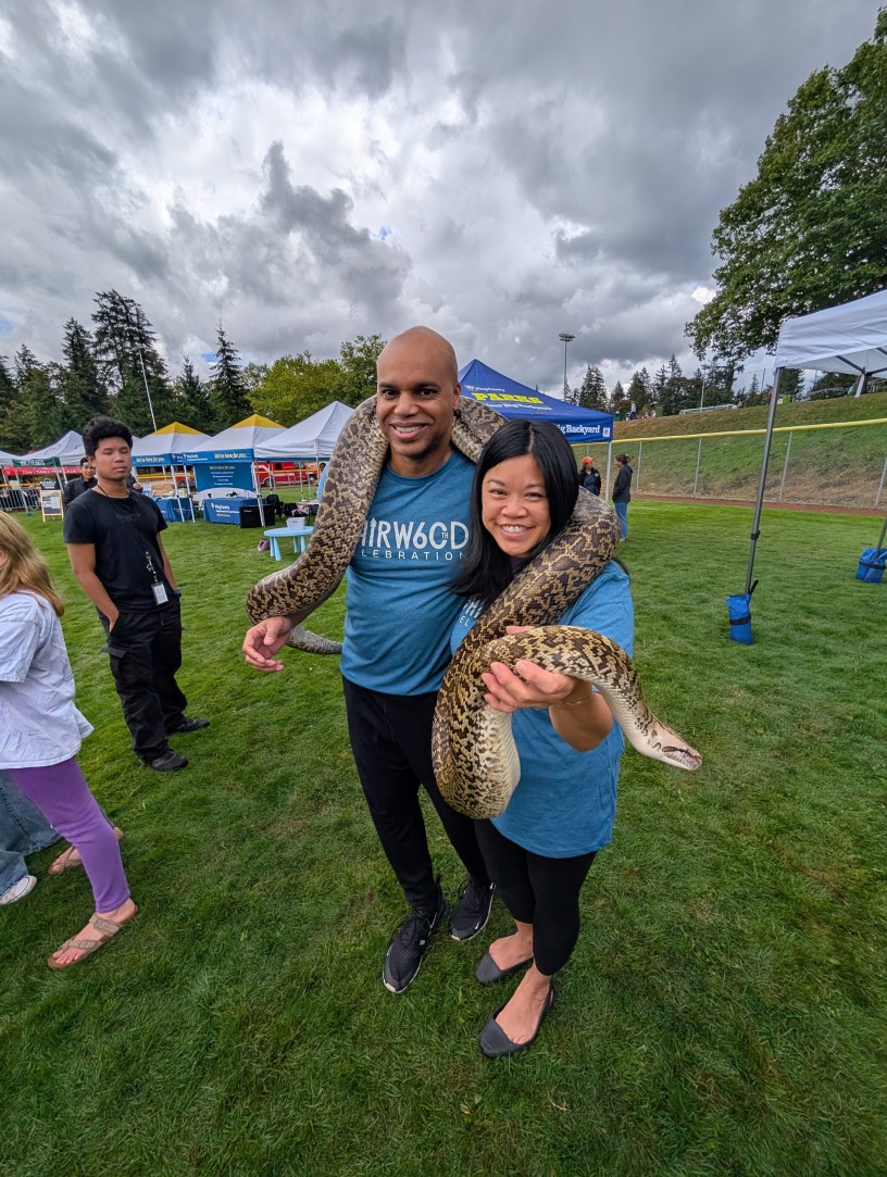 Two adults pose wearing t-shirts celebrating Fairwood's 60th anniversary pose for a picture with a large snake around their shoulders. They are standing in an open fields, and in the background are other people, tents, trees, and gloomy sky with gray clouds.
