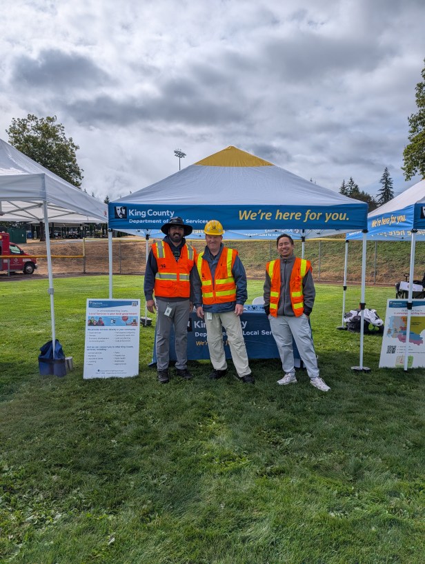 A trio of adults wearing orange vests pose for a picture at Fairwood's 60th Anniversary Celebration on Saturday, Aug. 29, 2026. They are standing in a grassy area and are next to an "A" board, with a department-branded tent, trees, and gray sky behind them.