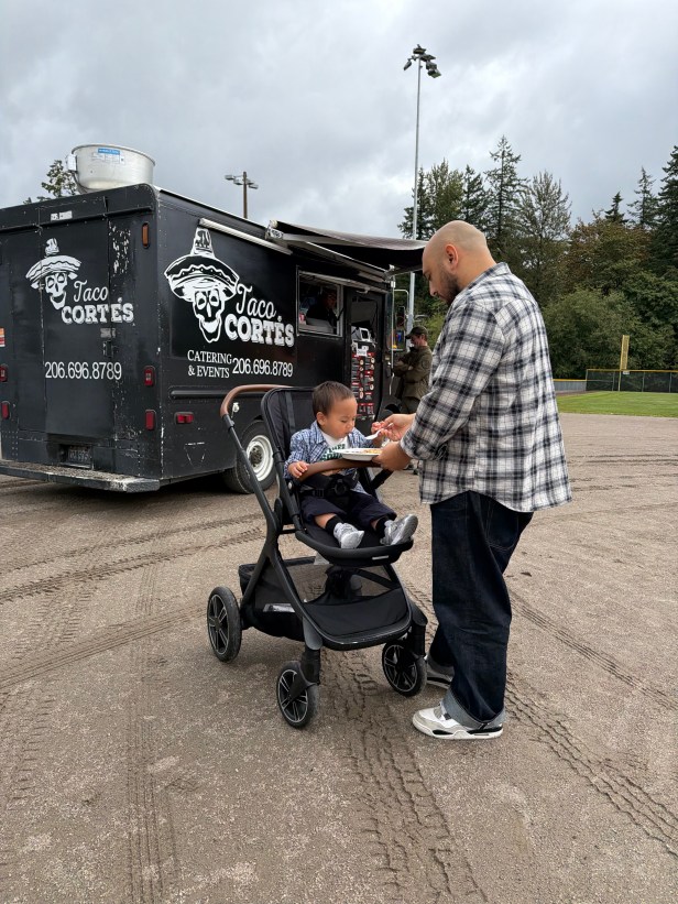 An adult feeds a young person in a stroller at Fairwood's 60th Anniversary Celebration on Saturday, Aug. 29, 2026. They are standing in the infield of a softball field, and in the background is a mobile food truck, trees, and gray sky.