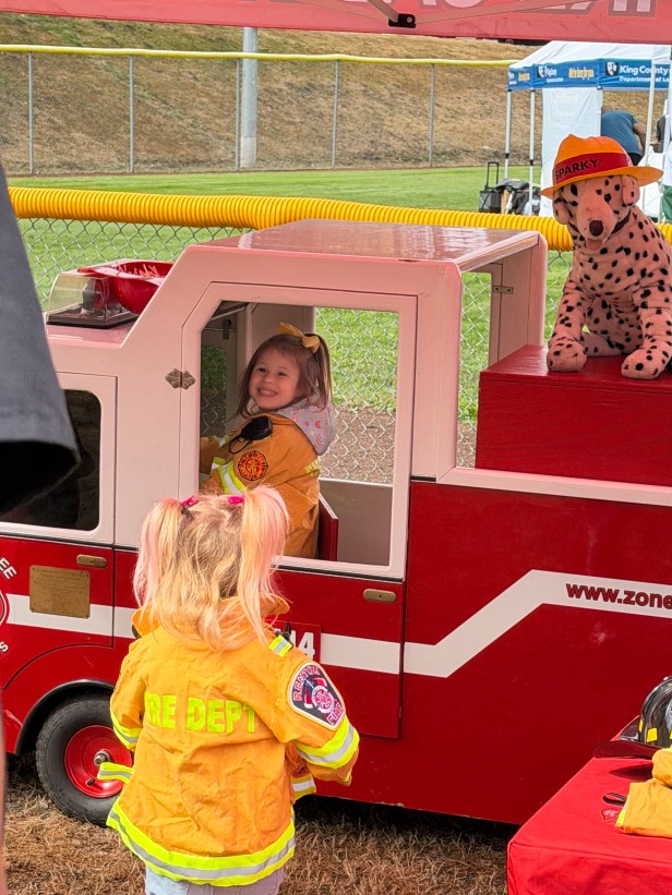 A young person smiles sitting behind the wheel of a miniature fire truck at A young person speaks to an adult table at the Zone Three Fire Cadets table at Fairwood's 60th Anniversary Celebration on Saturday, Aug. 29, 2026 at Petrovistky Park. On top of the truck is a stuffed animal of a Dalmatian dog, and a young person is in the foreground.