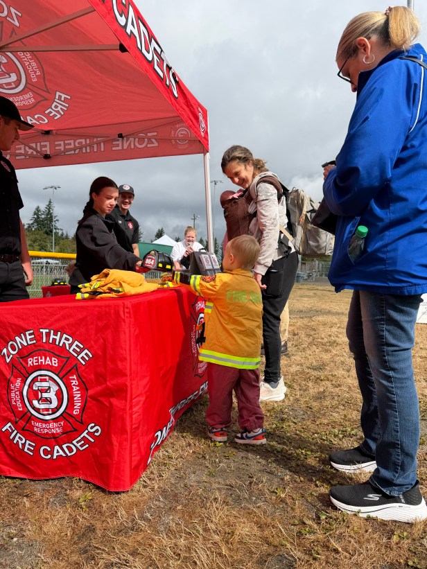 A young person speaks to an adult table at the Zone Three Fire Cadets table at Fairwood's 60th Anniversary Celebration on Saturday, Aug. 29, 2026 at Petrovistky Park. In the background in a fire truck, and they are in a grassy area. Another adult is in the foreground.