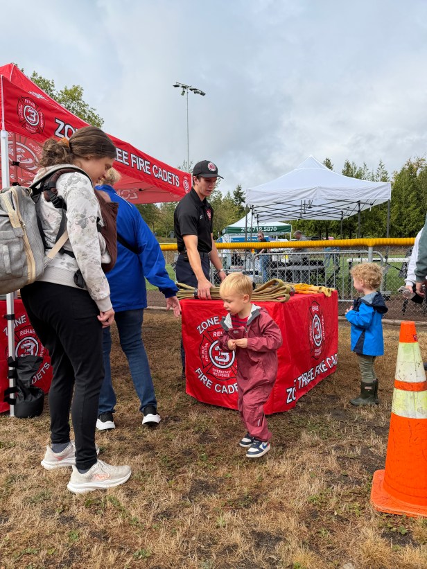 A young person speaks to an adult table at the Zone Three Fire Cadets table at Fairwood's 60th Anniversary Celebration on Saturday, Aug. 29, 2026 at Petrovistky Park. In the background in a fire truck, and they are in a grassy area. Another adult is in the foreground, along with a cone.
