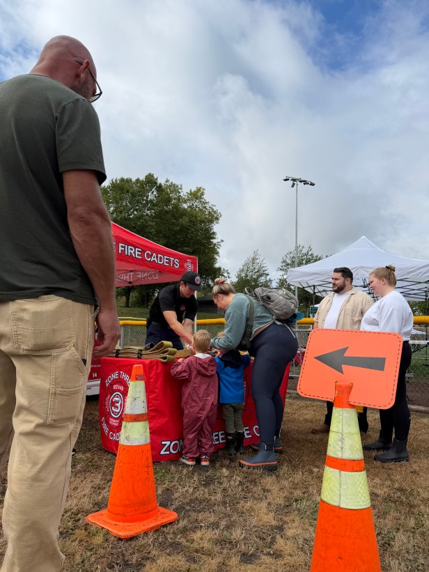 Two young people look at a table hosted by the Zone Three Fire Cadets at Fairwood's 60th Anniversary Celebration on Saturday, Aug. 29, 2026 at Petrovistky Park. They are standing next to an adult with two adults nearby, and another adult in the foreground, along with orange cones and signage.