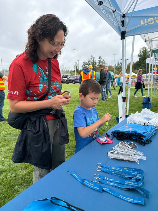 An adult and young person visit a table under a tent at a community festival. The table is covered, and on top are promotional materials including department-branded key chains, pens, stickers and drawstring backpacks. In the background is grass and other people.