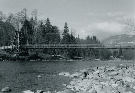 A black and white photo of the previous Baring Bridge in northeast King County. A wooden suspension bridge cross across the Middle For Snoqualmie River. In the foreground is a rocky river's edge, while on the other side is forested shoreline. In the background is a mountain with snow and clouds.