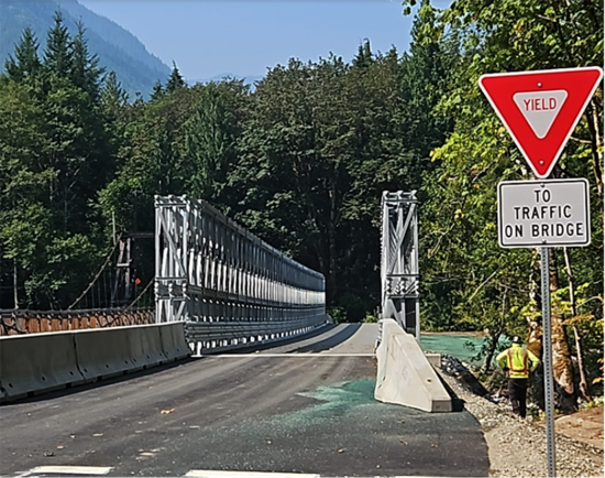 Image of a temporary bridge in Baring, Washington. The bridge is an Acrow Bridge. At the forefront is a yield sign with a sign that reads, "To Traffic on Bridge," next to a paved road with concrete barriers leading up to the bridge. In the background are trees and blue sky.