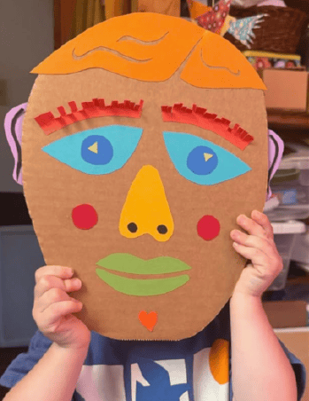 young child holds up painted cardboard mask covering face